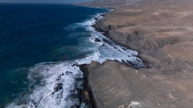  Kuvvetli dalgalarla kayalıklara çarpan kayalık kıyı şeridi berrak bir gökyüzü altında, derin mavi okyanus ve çorak kara arasındaki zıtlığı gözler önüne seriyor..
