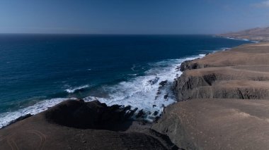  Kuvvetli dalgalarla kayalıklara çarpan kayalık kıyı şeridi berrak bir gökyüzü altında, derin mavi okyanus ve çorak kara arasındaki zıtlığı gözler önüne seriyor..