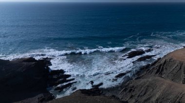  Kuvvetli dalgalarla kayalıklara çarpan kayalık kıyı şeridi berrak bir gökyüzü altında, derin mavi okyanus ve çorak kara arasındaki zıtlığı gözler önüne seriyor..
