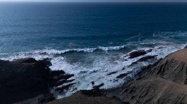  Kuvvetli dalgalarla kayalıklara çarpan kayalık kıyı şeridi berrak bir gökyüzü altında, derin mavi okyanus ve çorak kara arasındaki zıtlığı gözler önüne seriyor..