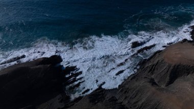  Kuvvetli dalgalarla kayalıklara çarpan kayalık kıyı şeridi berrak bir gökyüzü altında, derin mavi okyanus ve çorak kara arasındaki zıtlığı gözler önüne seriyor..