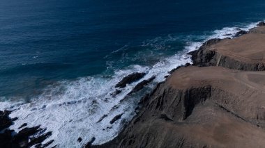  Kuvvetli dalgalarla kayalıklara çarpan kayalık kıyı şeridi berrak bir gökyüzü altında, derin mavi okyanus ve çorak kara arasındaki zıtlığı gözler önüne seriyor..