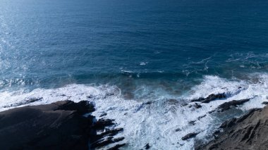  Kuvvetli dalgalarla kayalıklara çarpan kayalık kıyı şeridi berrak bir gökyüzü altında, derin mavi okyanus ve çorak kara arasındaki zıtlığı gözler önüne seriyor..