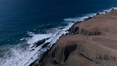  Kuvvetli dalgalarla kayalıklara çarpan kayalık kıyı şeridi berrak bir gökyüzü altında, derin mavi okyanus ve çorak kara arasındaki zıtlığı gözler önüne seriyor..