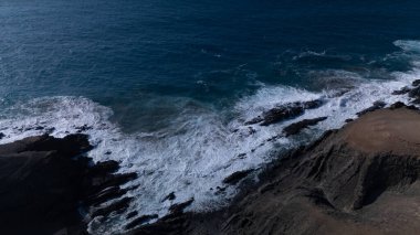  Kuvvetli dalgalarla kayalıklara çarpan kayalık kıyı şeridi berrak bir gökyüzü altında, derin mavi okyanus ve çorak kara arasındaki zıtlığı gözler önüne seriyor..