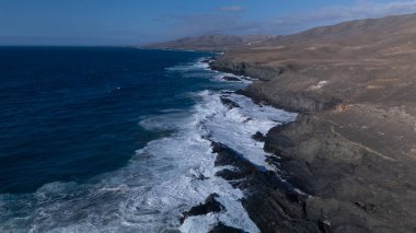 Kuvvetli dalgalarla kayalıklara çarpan kayalık kıyı şeridi berrak bir gökyüzü altında, derin mavi okyanus ve çorak kara arasındaki zıtlığı gözler önüne seriyor..