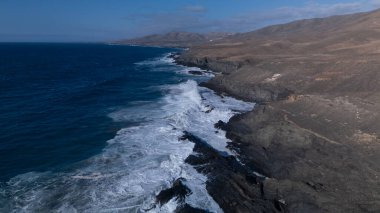 Kuvvetli dalgalarla kayalıklara çarpan kayalık kıyı şeridi berrak bir gökyüzü altında, derin mavi okyanus ve çorak kara arasındaki zıtlığı gözler önüne seriyor..