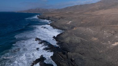 Kuvvetli dalgalarla kayalıklara çarpan kayalık kıyı şeridi berrak bir gökyüzü altında, derin mavi okyanus ve çorak kara arasındaki zıtlığı gözler önüne seriyor..