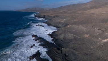 Kuvvetli dalgalarla kayalıklara çarpan kayalık kıyı şeridi berrak bir gökyüzü altında, derin mavi okyanus ve çorak kara arasındaki zıtlığı gözler önüne seriyor..