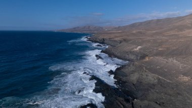 Kuvvetli dalgalarla kayalıklara çarpan kayalık kıyı şeridi berrak bir gökyüzü altında, derin mavi okyanus ve çorak kara arasındaki zıtlığı gözler önüne seriyor..