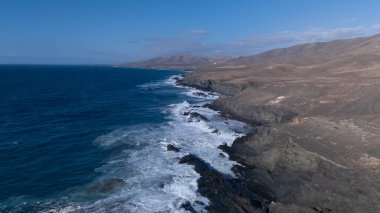 Kuvvetli dalgalarla kayalıklara çarpan kayalık kıyı şeridi berrak bir gökyüzü altında, derin mavi okyanus ve çorak kara arasındaki zıtlığı gözler önüne seriyor..