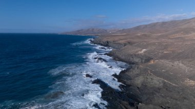 Kuvvetli dalgalarla kayalıklara çarpan kayalık kıyı şeridi berrak bir gökyüzü altında, derin mavi okyanus ve çorak kara arasındaki zıtlığı gözler önüne seriyor..