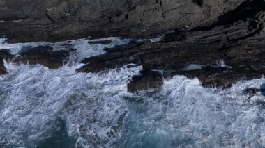 Kuvvetli dalgalarla kayalıklara çarpan kayalık kıyı şeridi berrak bir gökyüzü altında, derin mavi okyanus ve çorak kara arasındaki zıtlığı gözler önüne seriyor..