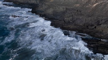Kuvvetli dalgalarla kayalıklara çarpan kayalık kıyı şeridi berrak bir gökyüzü altında, derin mavi okyanus ve çorak kara arasındaki zıtlığı gözler önüne seriyor..