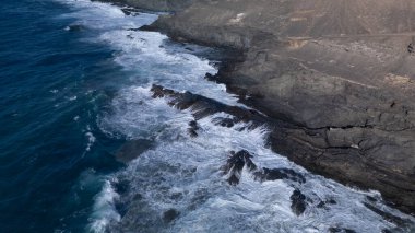 Kuvvetli dalgalarla kayalıklara çarpan kayalık kıyı şeridi berrak bir gökyüzü altında, derin mavi okyanus ve çorak kara arasındaki zıtlığı gözler önüne seriyor..