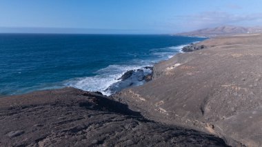 Kuvvetli dalgalarla kayalıklara çarpan kayalık kıyı şeridi berrak bir gökyüzü altında, derin mavi okyanus ve çorak kara arasındaki zıtlığı gözler önüne seriyor..