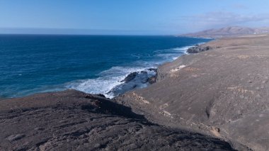 Kuvvetli dalgalarla kayalıklara çarpan kayalık kıyı şeridi berrak bir gökyüzü altında, derin mavi okyanus ve çorak kara arasındaki zıtlığı gözler önüne seriyor..