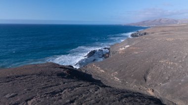 Kuvvetli dalgalarla kayalıklara çarpan kayalık kıyı şeridi berrak bir gökyüzü altında, derin mavi okyanus ve çorak kara arasındaki zıtlığı gözler önüne seriyor..