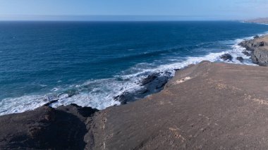 Kuvvetli dalgalarla kayalıklara çarpan kayalık kıyı şeridi berrak bir gökyüzü altında, derin mavi okyanus ve çorak kara arasındaki zıtlığı gözler önüne seriyor..