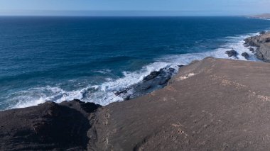 Kuvvetli dalgalarla kayalıklara çarpan kayalık kıyı şeridi berrak bir gökyüzü altında, derin mavi okyanus ve çorak kara arasındaki zıtlığı gözler önüne seriyor..