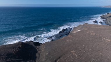 Kuvvetli dalgalarla kayalıklara çarpan kayalık kıyı şeridi berrak bir gökyüzü altında, derin mavi okyanus ve çorak kara arasındaki zıtlığı gözler önüne seriyor..