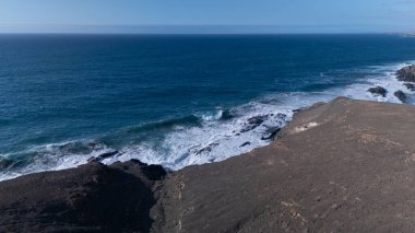 Kuvvetli dalgalarla kayalıklara çarpan kayalık kıyı şeridi berrak bir gökyüzü altında, derin mavi okyanus ve çorak kara arasındaki zıtlığı gözler önüne seriyor..