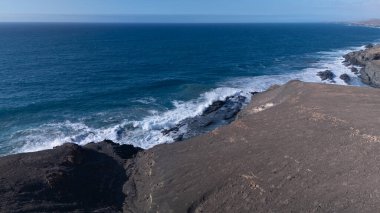 Kuvvetli dalgalarla kayalıklara çarpan kayalık kıyı şeridi berrak bir gökyüzü altında, derin mavi okyanus ve çorak kara arasındaki zıtlığı gözler önüne seriyor..