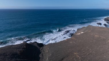 Kuvvetli dalgalarla kayalıklara çarpan kayalık kıyı şeridi berrak bir gökyüzü altında, derin mavi okyanus ve çorak kara arasındaki zıtlığı gözler önüne seriyor..