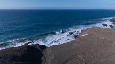 Kuvvetli dalgalarla kayalıklara çarpan kayalık kıyı şeridi berrak bir gökyüzü altında, derin mavi okyanus ve çorak kara arasındaki zıtlığı gözler önüne seriyor..
