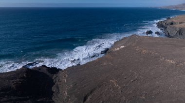 Kuvvetli dalgalarla kayalıklara çarpan kayalık kıyı şeridi berrak bir gökyüzü altında, derin mavi okyanus ve çorak kara arasındaki zıtlığı gözler önüne seriyor..
