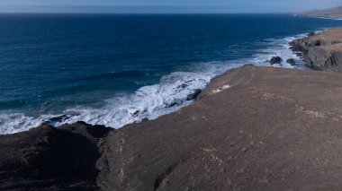 Kuvvetli dalgalarla kayalıklara çarpan kayalık kıyı şeridi berrak bir gökyüzü altında, derin mavi okyanus ve çorak kara arasındaki zıtlığı gözler önüne seriyor..