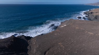 Kuvvetli dalgalarla kayalıklara çarpan kayalık kıyı şeridi berrak bir gökyüzü altında, derin mavi okyanus ve çorak kara arasındaki zıtlığı gözler önüne seriyor..