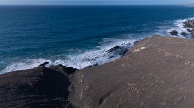  Kuvvetli dalgalarla kayalıklara çarpan kayalık kıyı şeridi berrak bir gökyüzü altında, derin mavi okyanus ve çorak kara arasındaki zıtlığı gözler önüne seriyor..