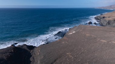 Kuvvetli dalgalarla kayalıklara çarpan kayalık kıyı şeridi berrak bir gökyüzü altında, derin mavi okyanus ve çorak kara arasındaki zıtlığı gözler önüne seriyor..