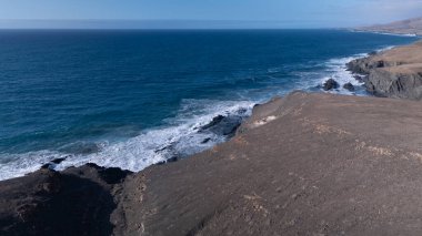 Kuvvetli dalgalarla kayalıklara çarpan kayalık kıyı şeridi berrak bir gökyüzü altında, derin mavi okyanus ve çorak kara arasındaki zıtlığı gözler önüne seriyor..
