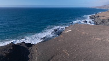 Kuvvetli dalgalarla kayalıklara çarpan kayalık kıyı şeridi berrak bir gökyüzü altında, derin mavi okyanus ve çorak kara arasındaki zıtlığı gözler önüne seriyor..