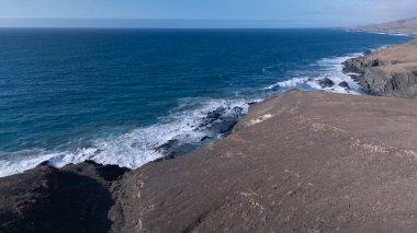 Kuvvetli dalgalarla kayalıklara çarpan kayalık kıyı şeridi berrak bir gökyüzü altında, derin mavi okyanus ve çorak kara arasındaki zıtlığı gözler önüne seriyor..