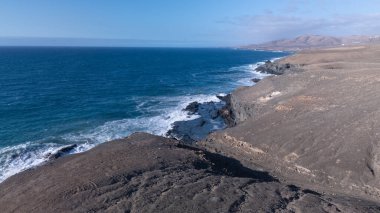 Kuvvetli dalgalarla kayalıklara çarpan kayalık kıyı şeridi berrak bir gökyüzü altında, derin mavi okyanus ve çorak kara arasındaki zıtlığı gözler önüne seriyor..