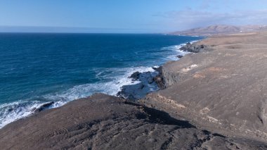 Kuvvetli dalgalarla kayalıklara çarpan kayalık kıyı şeridi berrak bir gökyüzü altında, derin mavi okyanus ve çorak kara arasındaki zıtlığı gözler önüne seriyor..