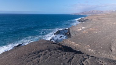 Kuvvetli dalgalarla kayalıklara çarpan kayalık kıyı şeridi berrak bir gökyüzü altında, derin mavi okyanus ve çorak kara arasındaki zıtlığı gözler önüne seriyor..