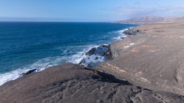 Kuvvetli dalgalarla kayalıklara çarpan kayalık kıyı şeridi berrak bir gökyüzü altında, derin mavi okyanus ve çorak kara arasındaki zıtlığı gözler önüne seriyor..