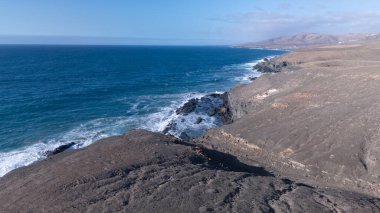 Kuvvetli dalgalarla kayalıklara çarpan kayalık kıyı şeridi berrak bir gökyüzü altında, derin mavi okyanus ve çorak kara arasındaki zıtlığı gözler önüne seriyor..