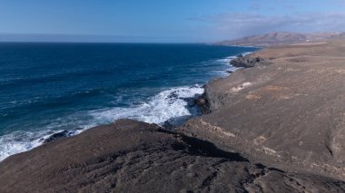 Kuvvetli dalgalarla kayalıklara çarpan kayalık kıyı şeridi berrak bir gökyüzü altında, derin mavi okyanus ve çorak kara arasındaki zıtlığı gözler önüne seriyor..
