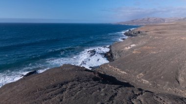 Kuvvetli dalgalarla kayalıklara çarpan kayalık kıyı şeridi berrak bir gökyüzü altında, derin mavi okyanus ve çorak kara arasındaki zıtlığı gözler önüne seriyor..