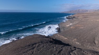 Kuvvetli dalgalarla kayalıklara çarpan kayalık kıyı şeridi berrak bir gökyüzü altında, derin mavi okyanus ve çorak kara arasındaki zıtlığı gözler önüne seriyor..