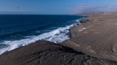 Kuvvetli dalgalarla kayalıklara çarpan kayalık kıyı şeridi berrak bir gökyüzü altında, derin mavi okyanus ve çorak kara arasındaki zıtlığı gözler önüne seriyor..