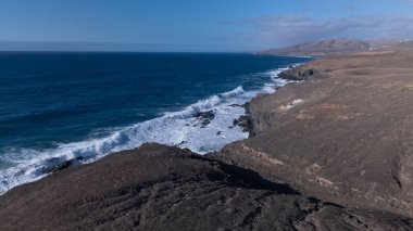 Kuvvetli dalgalarla kayalıklara çarpan kayalık kıyı şeridi berrak bir gökyüzü altında, derin mavi okyanus ve çorak kara arasındaki zıtlığı gözler önüne seriyor..