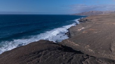 Kuvvetli dalgalarla kayalıklara çarpan kayalık kıyı şeridi berrak bir gökyüzü altında, derin mavi okyanus ve çorak kara arasındaki zıtlığı gözler önüne seriyor..