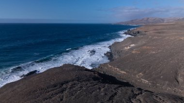 Kuvvetli dalgalarla kayalıklara çarpan kayalık kıyı şeridi berrak bir gökyüzü altında, derin mavi okyanus ve çorak kara arasındaki zıtlığı gözler önüne seriyor..