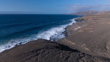 Kuvvetli dalgalarla kayalıklara çarpan kayalık kıyı şeridi berrak bir gökyüzü altında, derin mavi okyanus ve çorak kara arasındaki zıtlığı gözler önüne seriyor..